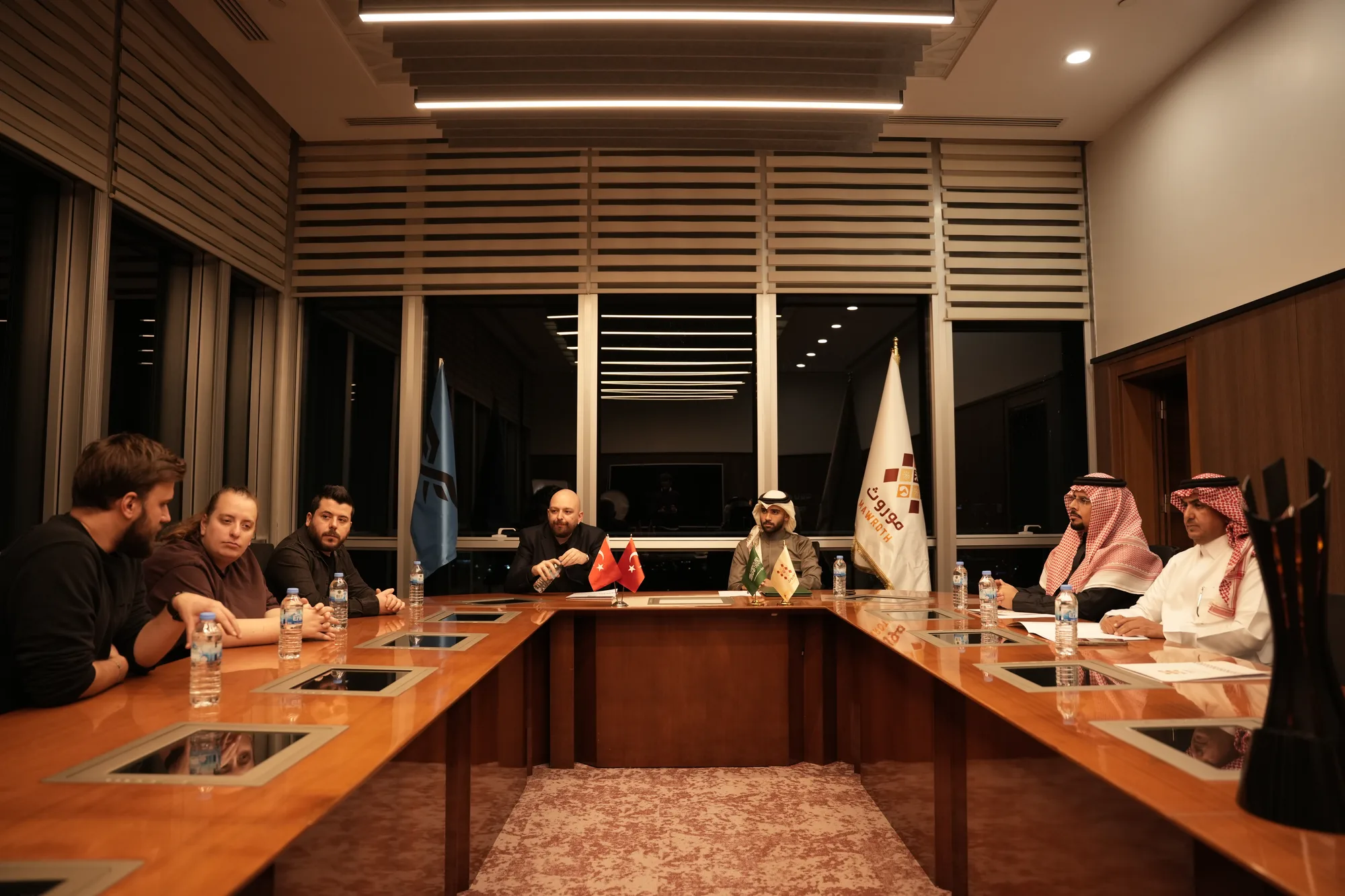 Both delegations seated at the full boardroom table under evening lighting, Mawroth and Fire Flux flags flanking the room