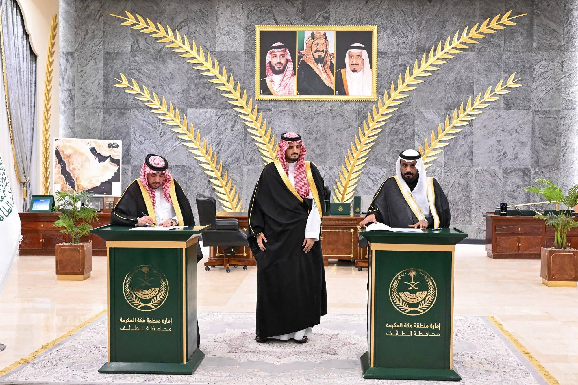 Three signatories at official podiums in Taif Governorate hall, overseen by HH Prince Fawaz bin Sultan, with royal portraits and gold palm-frond décor