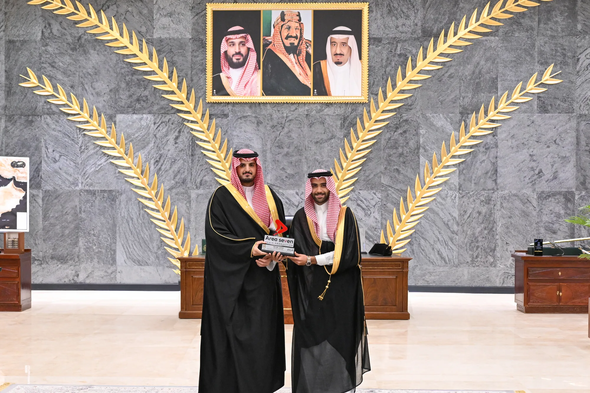 Two dignitaries exchanging a commemorative plaque in the Governor of Taif's reception hall, royal portraits and decorative gold palm fronds in the background