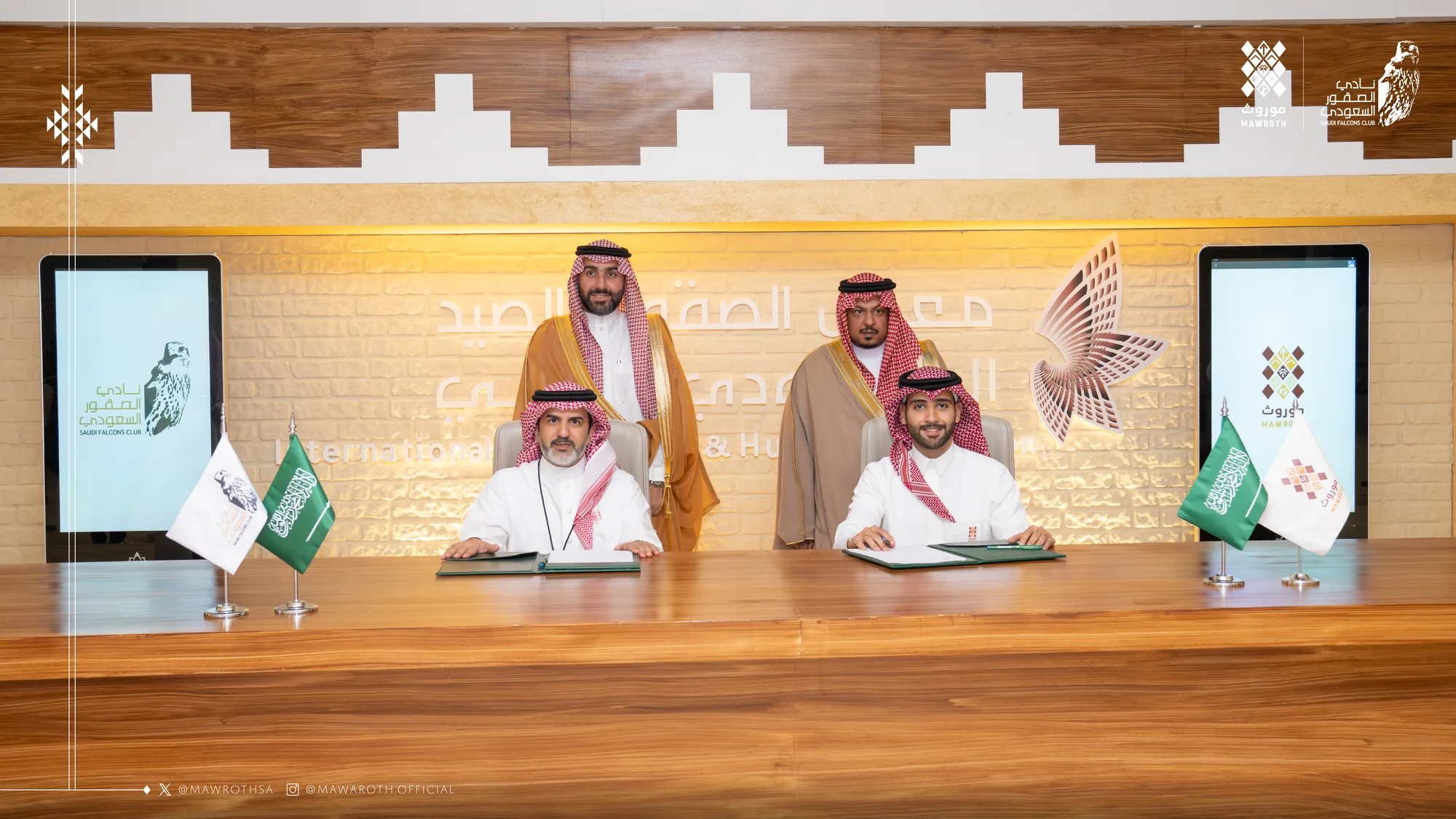 Two officials sign the Mawroth–Saudi Falcons Club cooperation agreement at the signing table, with two standing witnesses behind them