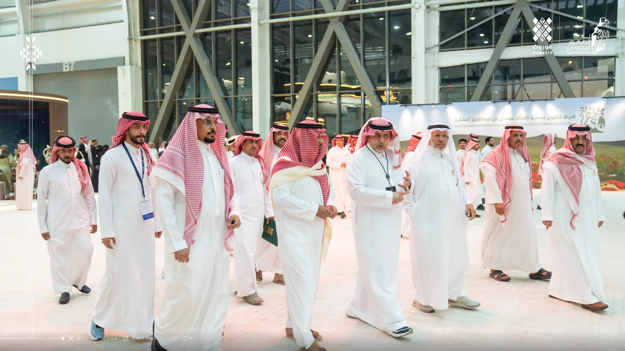 Large delegation of officials walking in formation through the exhibition entrance, Mawroth and Saudi Falcons Club logos overhead