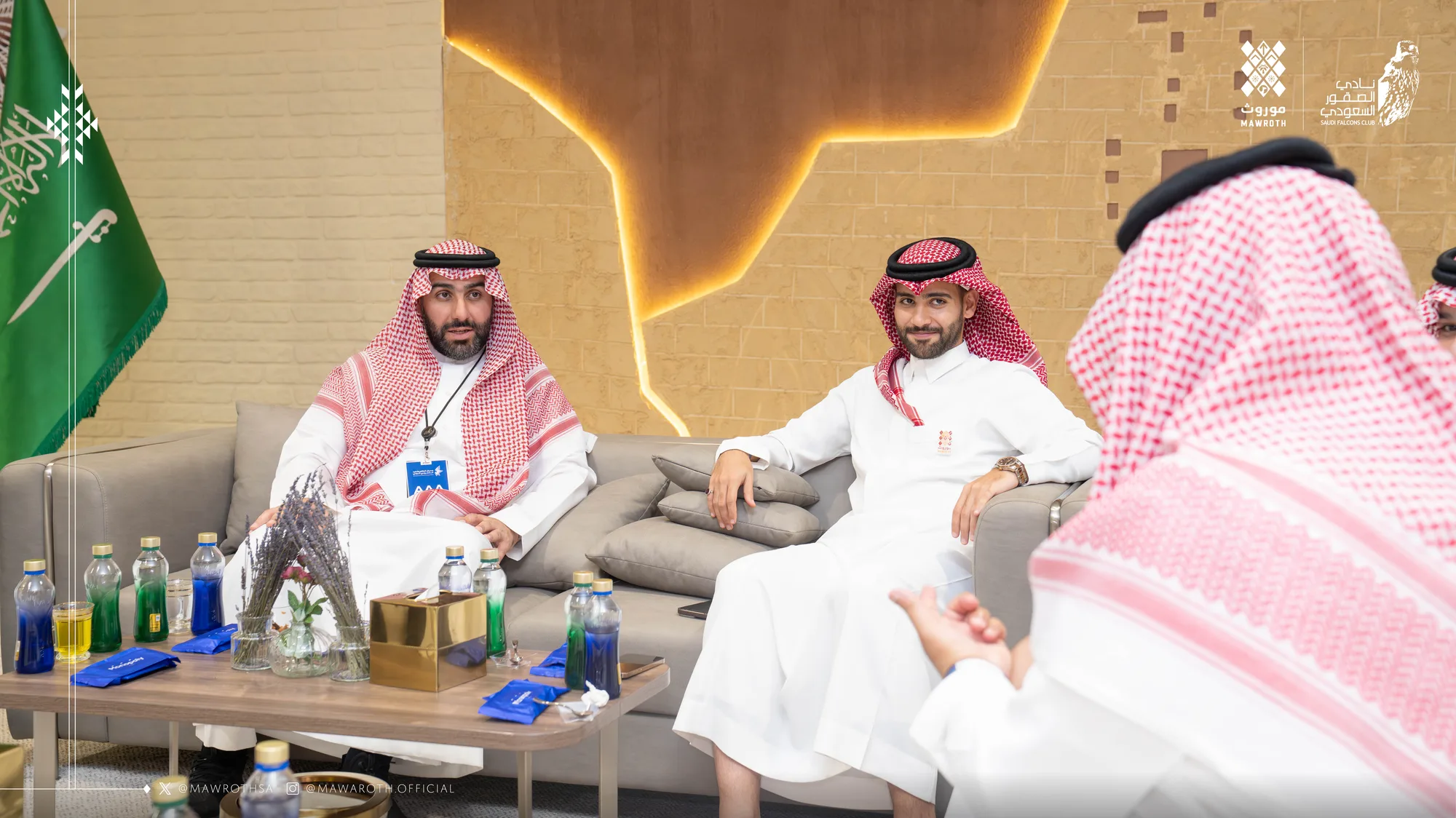 Two key principals in discussion across the meeting table at the exhibition lounge, Saudi flag in the background