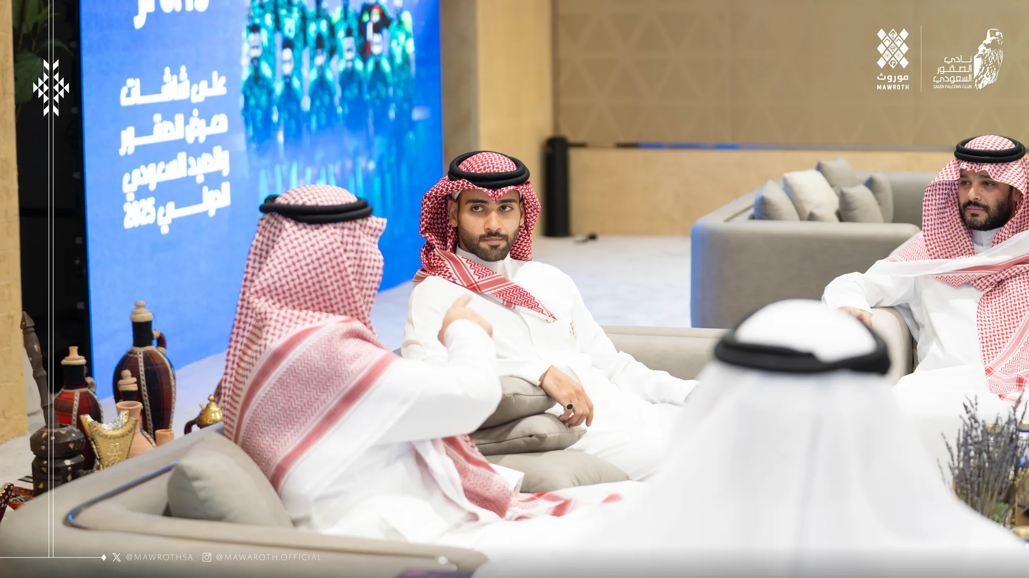 Three delegation members in relaxed discussion on couches at the exhibition meeting area, presentation screen visible