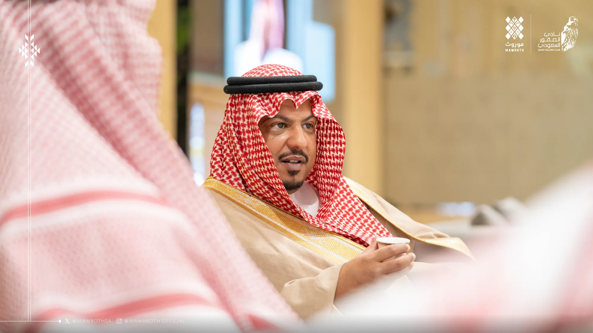 Senior dignitary in golden bisht engaged in conversation at the exhibition lounge, Mawroth and Saudi Falcons Club logos above