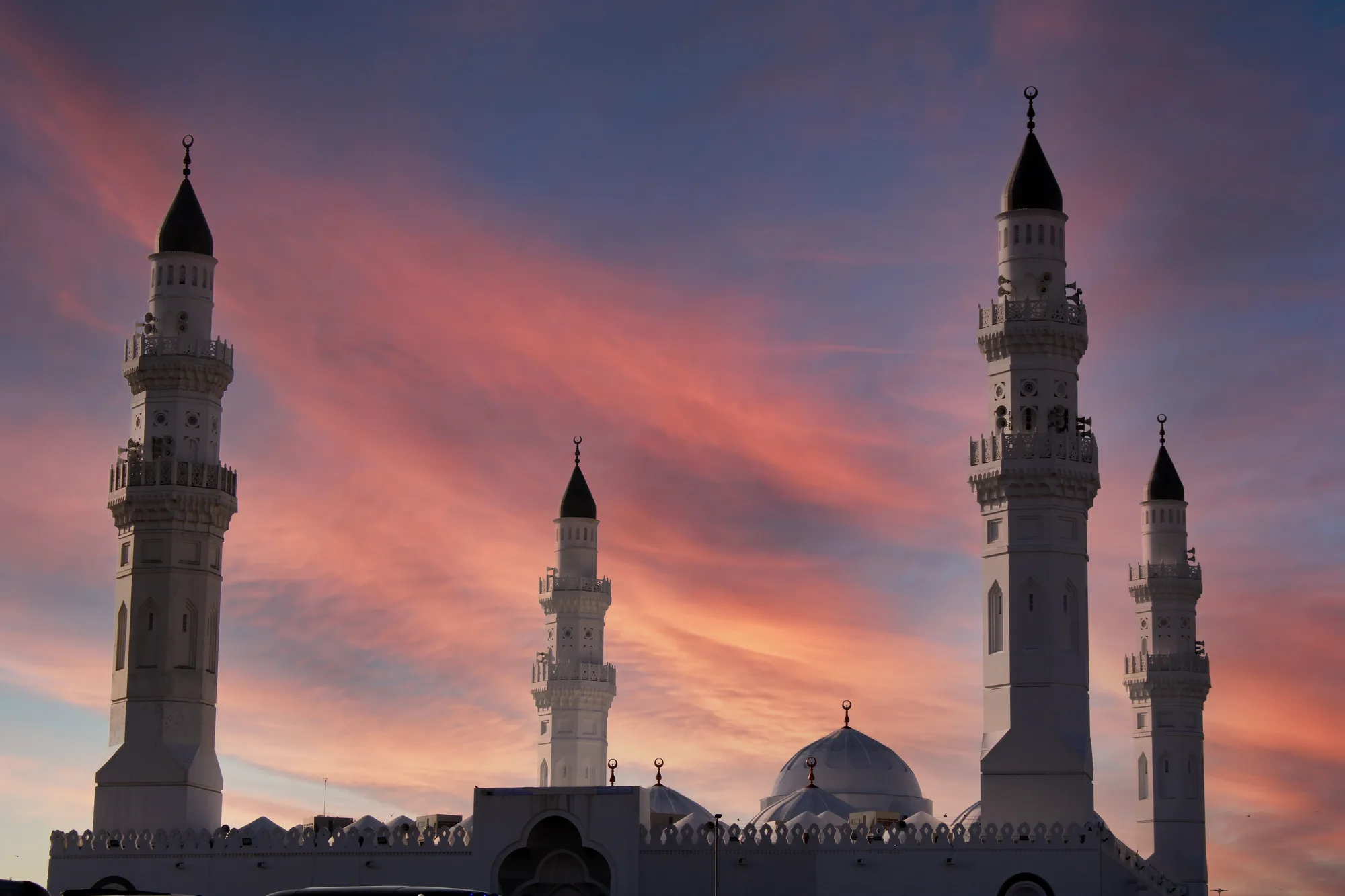Quba Mosque, Medina — four illuminated minarets silhouetted against a vivid crimson and violet sunset sky
