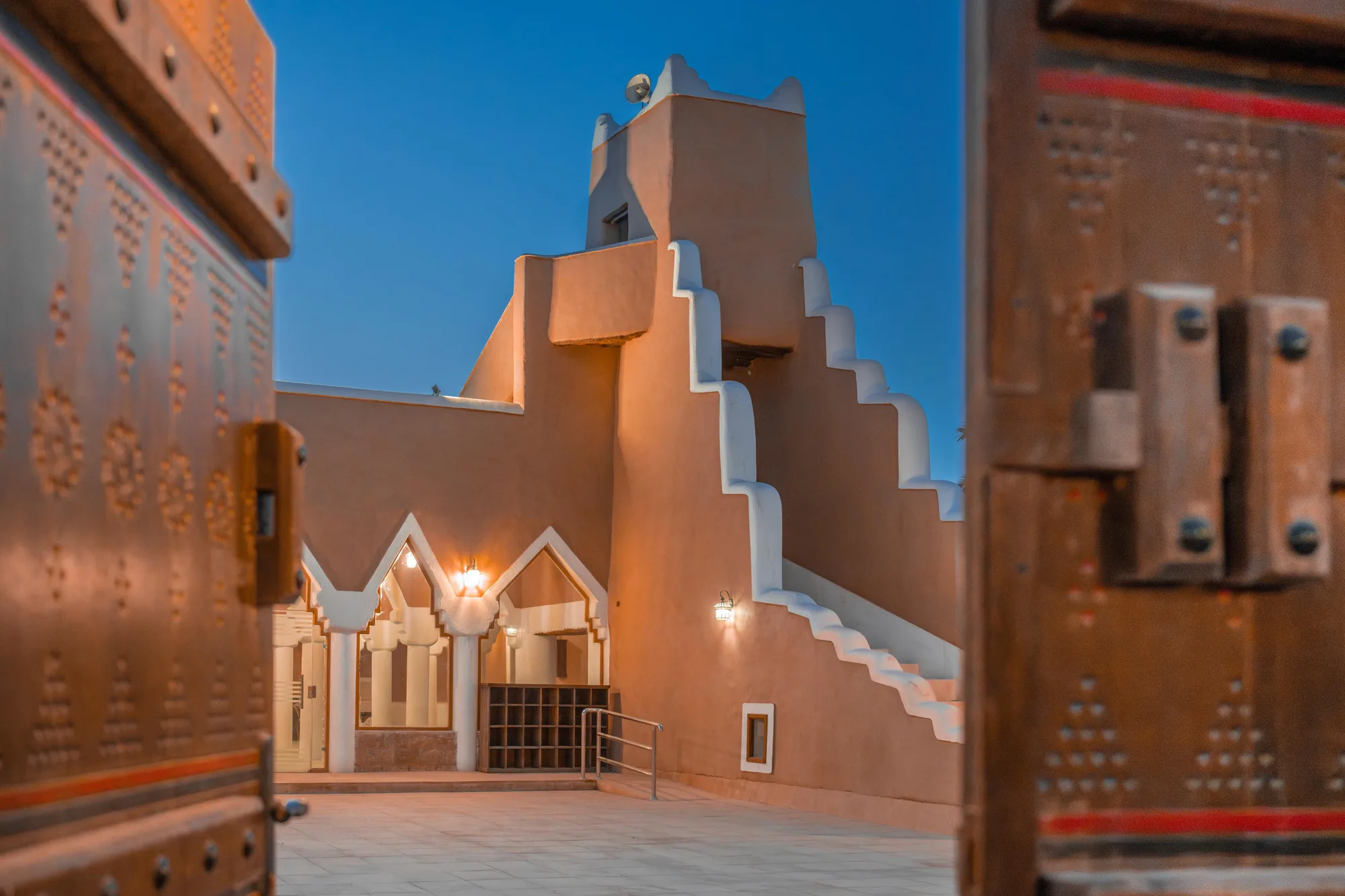 Restored Najdi mud-brick mosque at dusk, framed by ornate copper-studded doors swung open, stepped parapet and minaret glowing amber