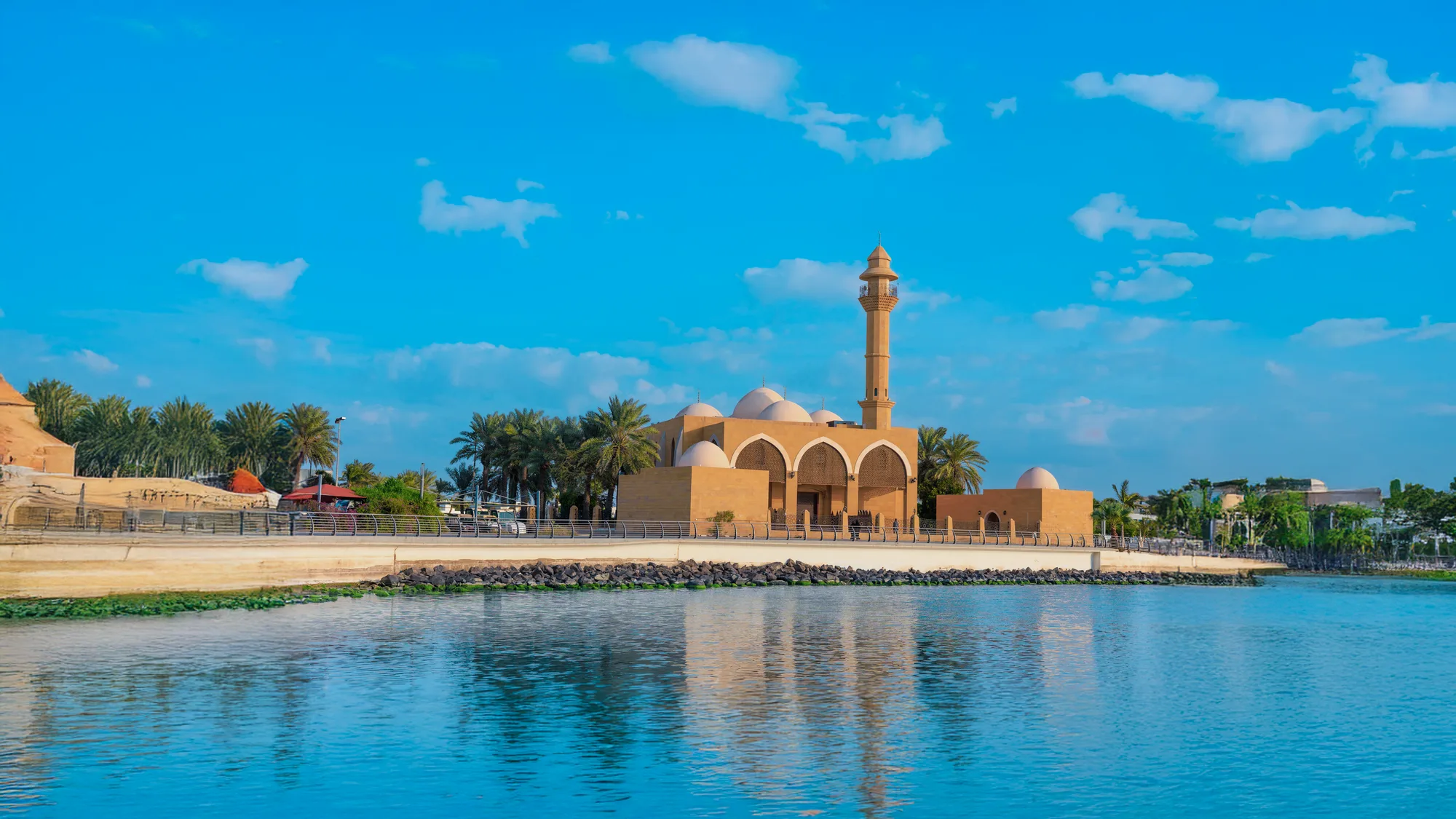 Sandy-toned waterfront mosque with arched portico and single minaret reflected in the still lagoon, Jeddah corniche