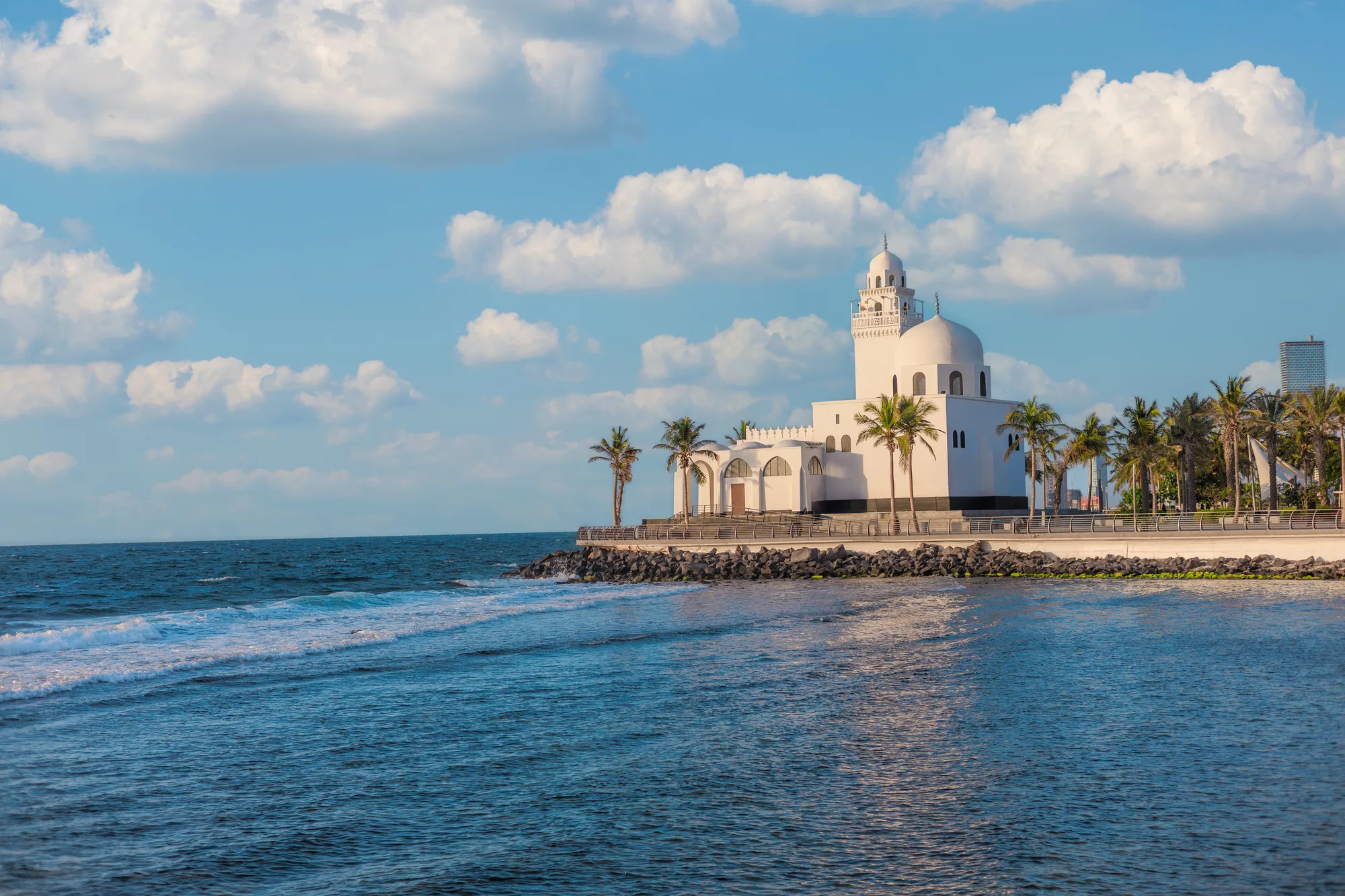 White domed Jeddah corniche mosque on a rocky promontory, flanked by palm trees, at golden hour with the Red Sea behind