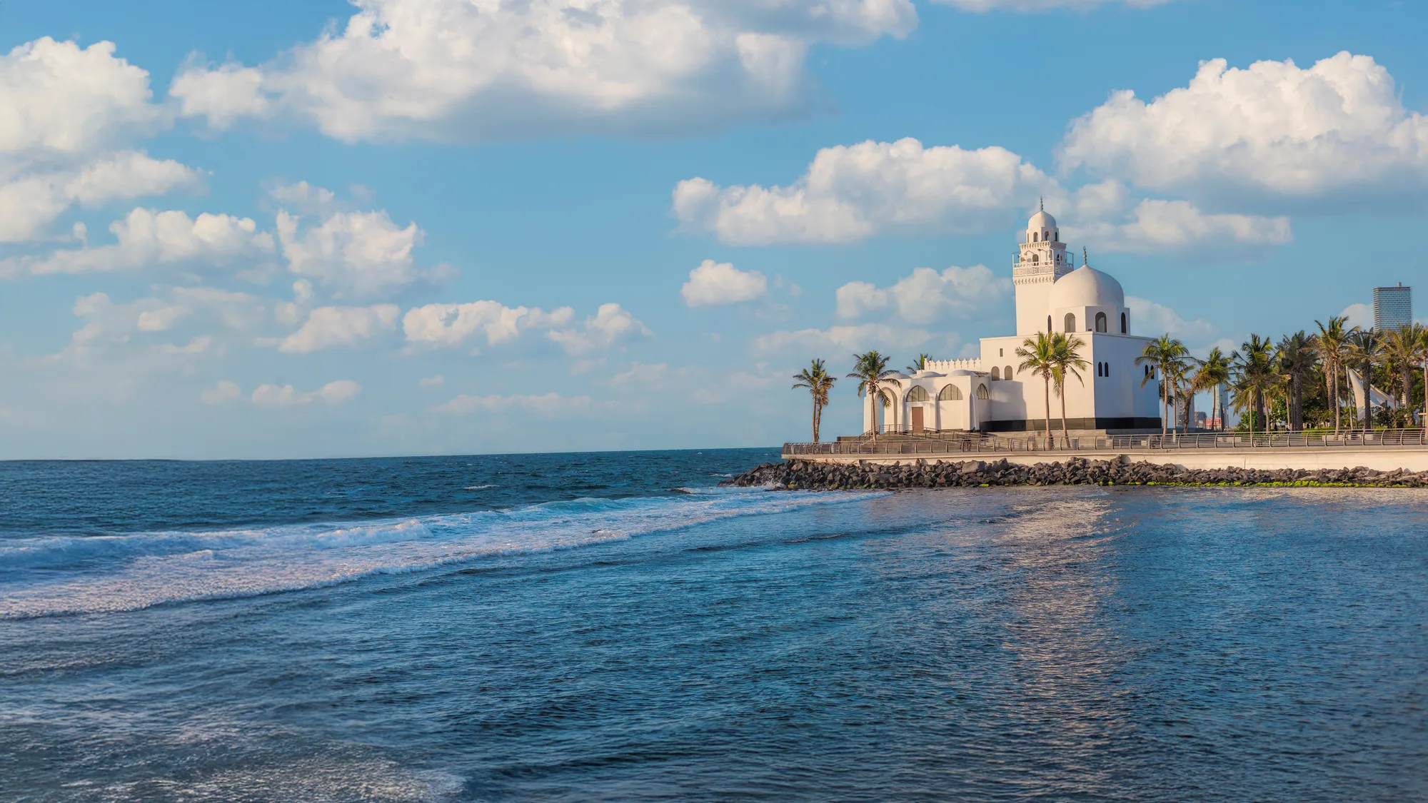 Wide-angle view of the white Jeddah waterfront mosque on its coastal breakwater, deep blue sky and ocean