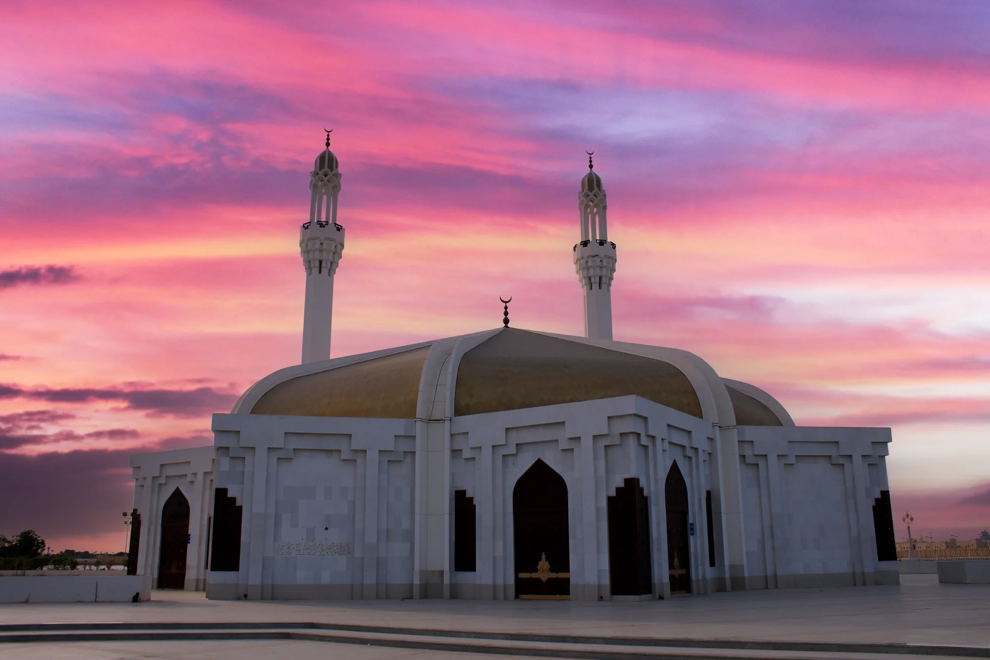 Hassan Any Mosque, Jeddah — two illuminated classical minarets rise against a blazing magenta sunset, mountain silhouette behind