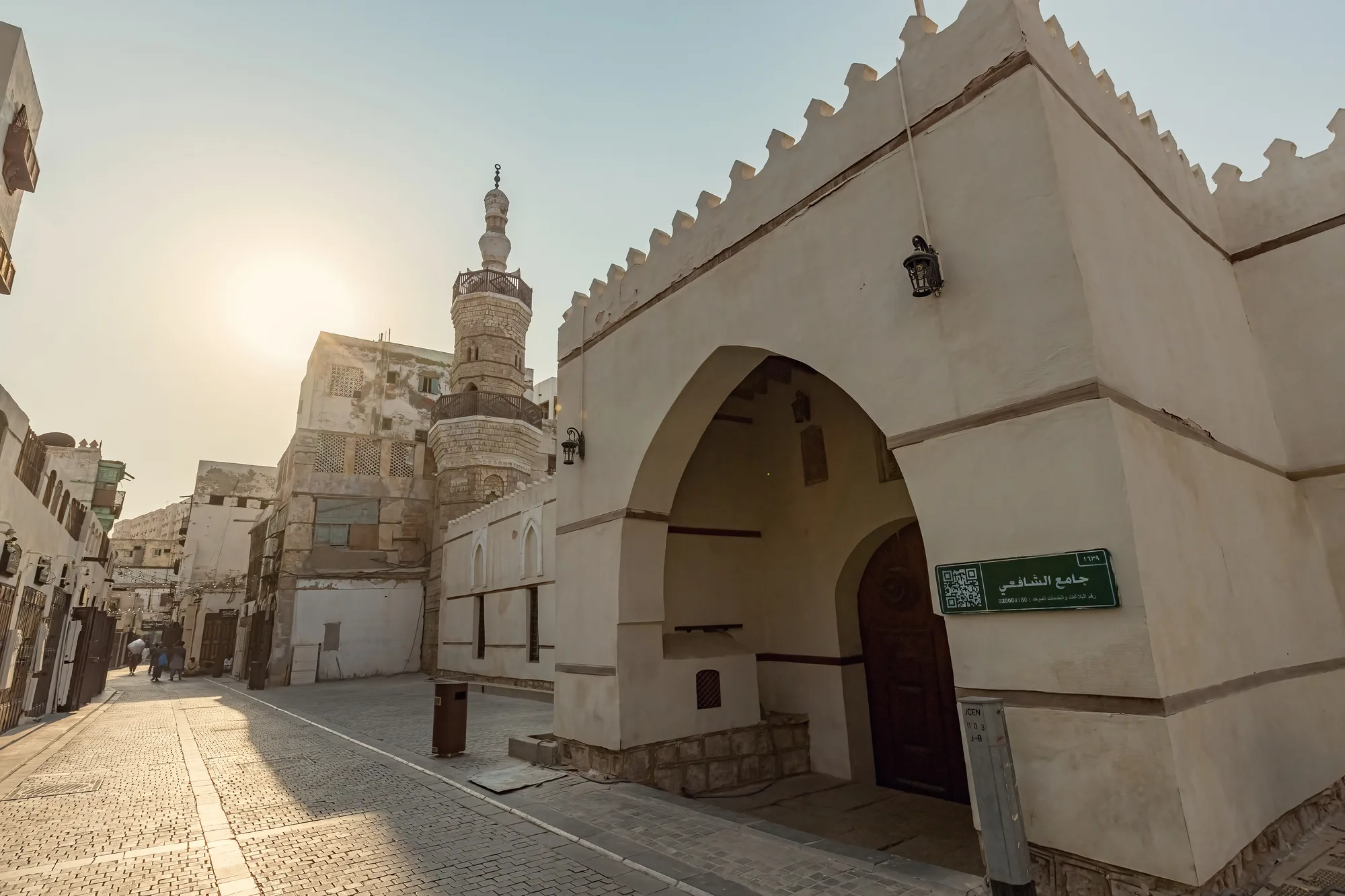Al-Shafei Mosque entrance arch in Al-Balad historic district, Jeddah — coral-stone minaret and green signboard, backlit by afternoon sun