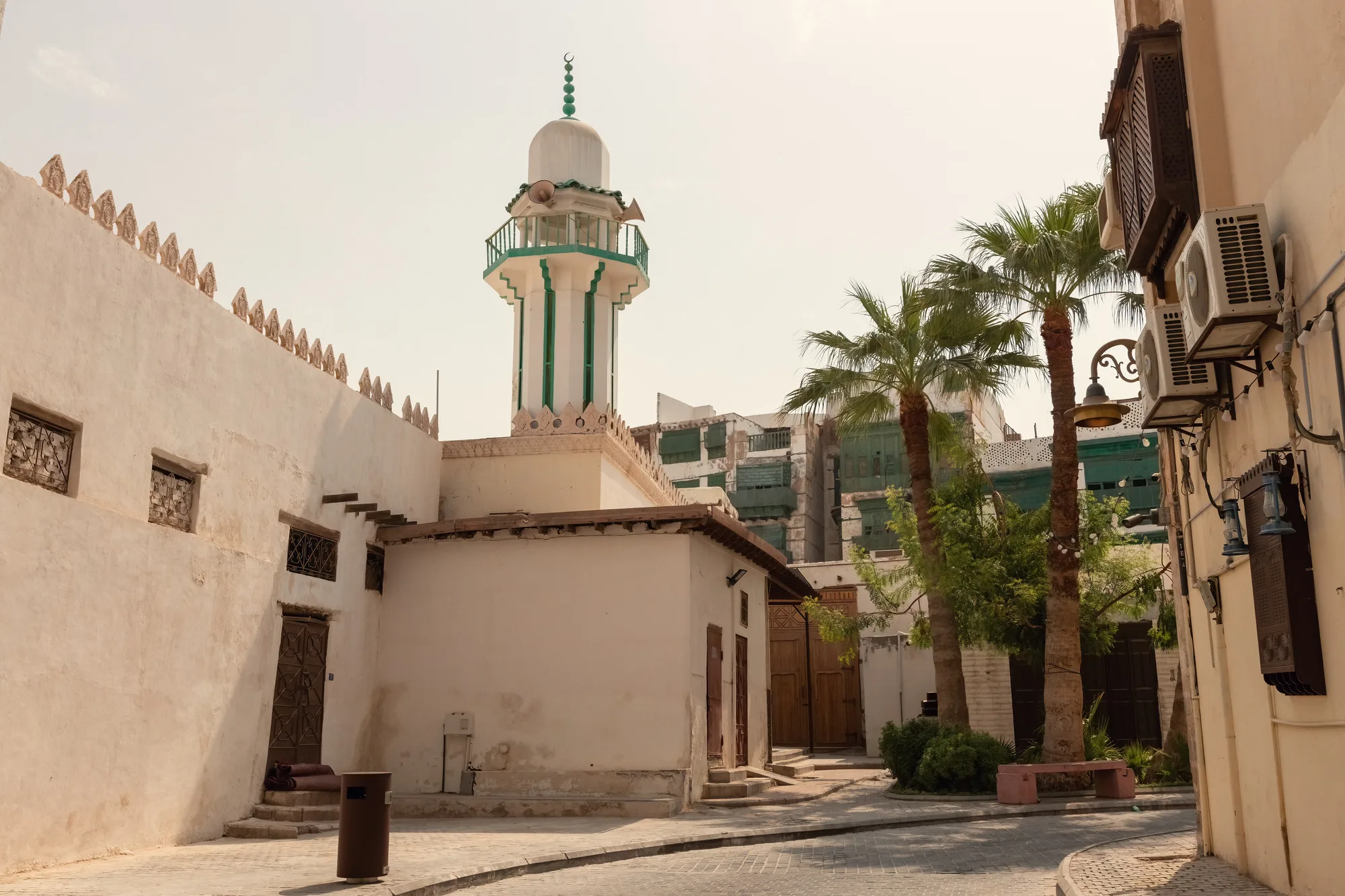 Historic Hanafi Mosque in Old Jeddah — white minaret with green stripe rising above a courtyard flanked by traditional Hijazi buildings and palm trees