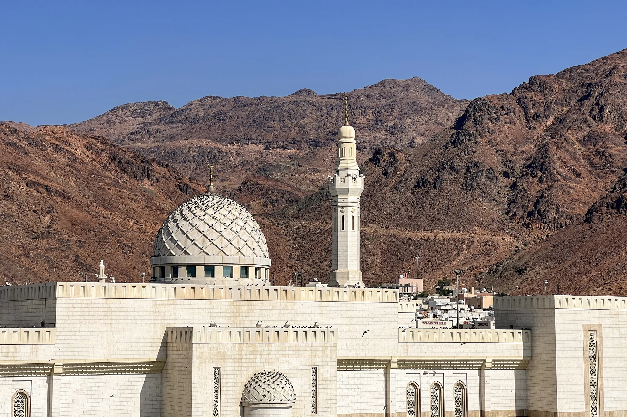 Sayyid al-Shuhada Mosque at the foot of Mount Uhud, Medina — ornate white dome and minaret against bare rocky mountains under clear blue sky