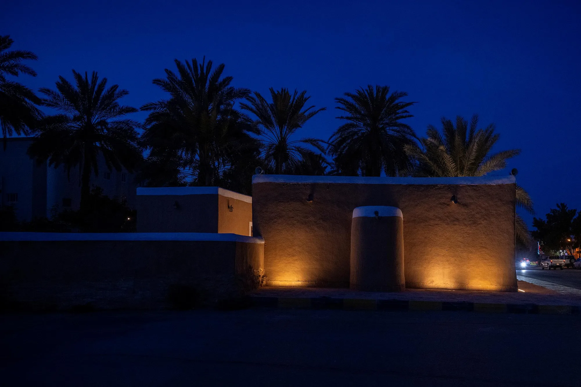Ancient mud-brick mosque perimeter wall illuminated by warm uplighting at night, silhouetted date palms against deep blue sky, Najd region