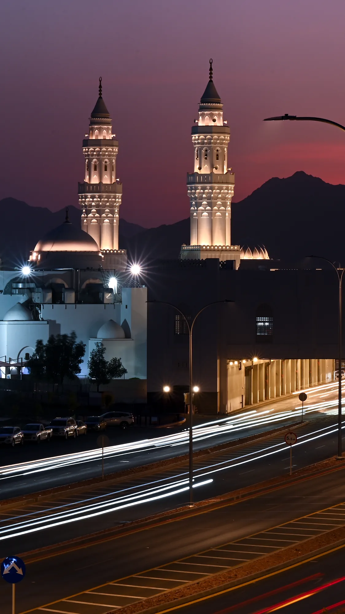 Twin illuminated minarets of a Saudi mosque rising against a deep purple dusk sky, long-exposure light trails from traffic below