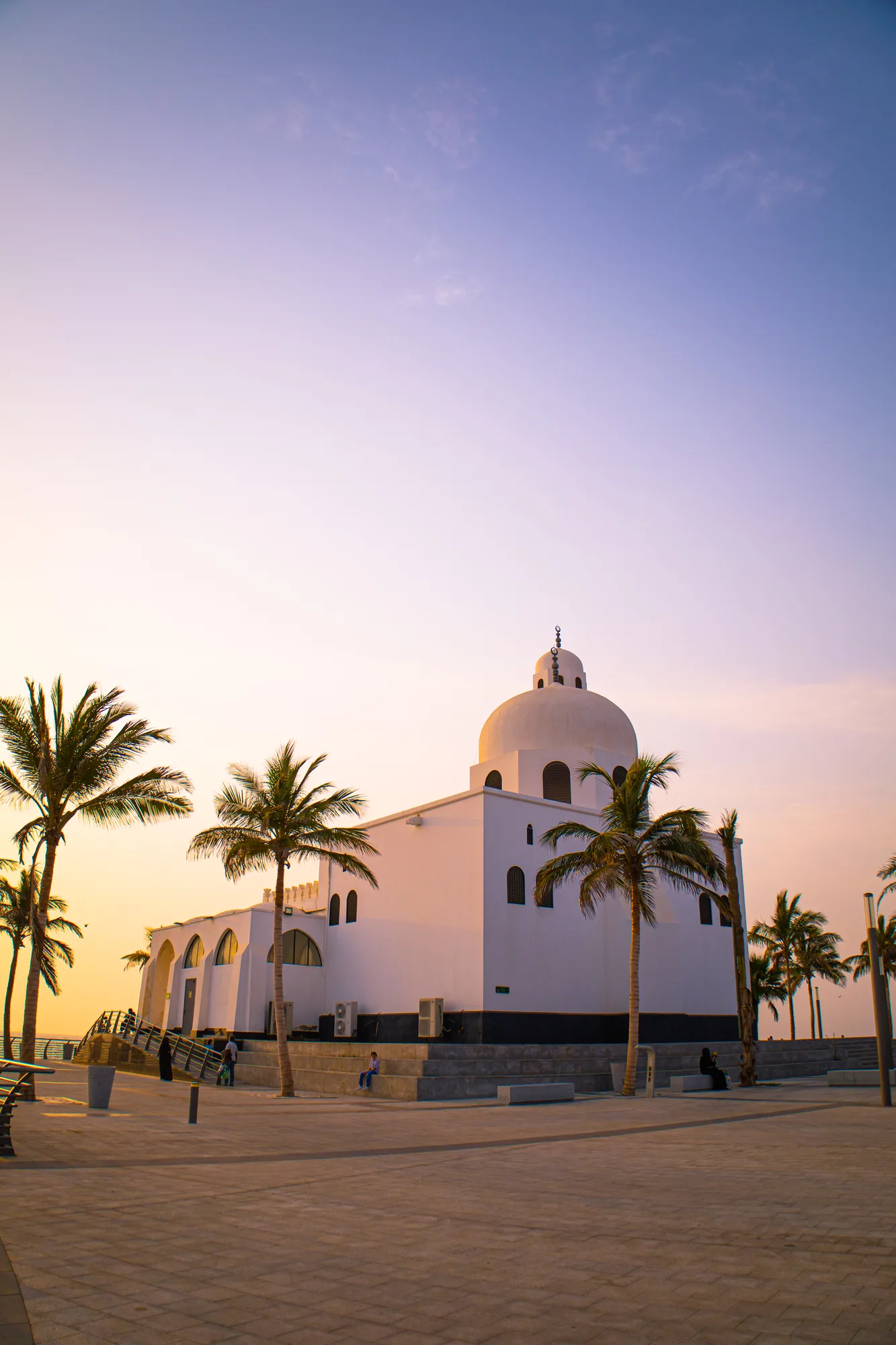 Jeddah Corniche Mosque at sunset — white domed structure beside tall palms, pastel sky of lavender and gold, promenade in foreground
