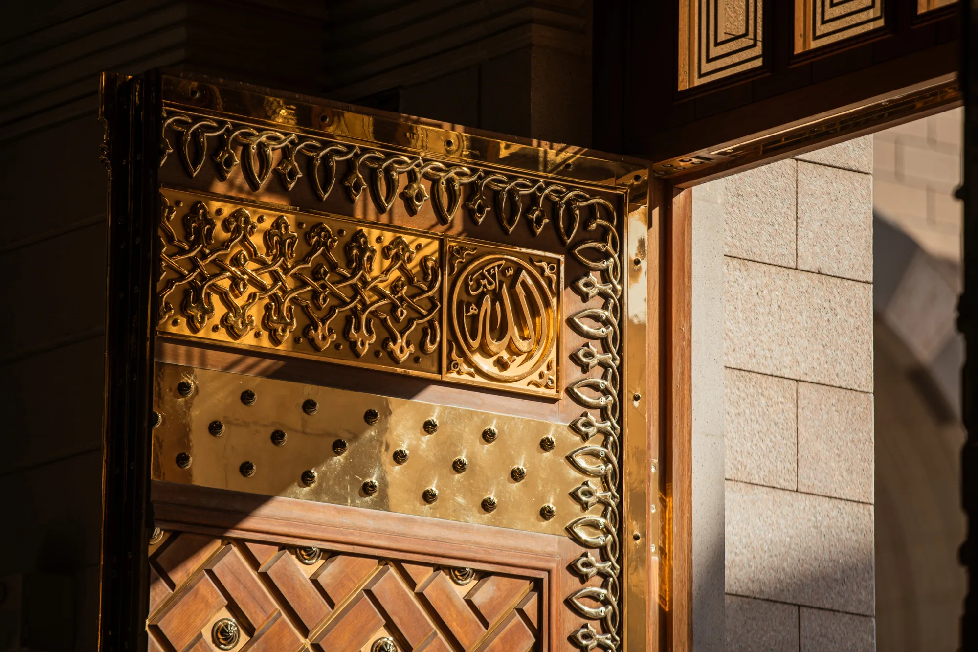 Close-up of an ornate gilded mosque door ajar — Arabic calligraphy, interlaced arabesque panels and brass studs catching warm sunlight