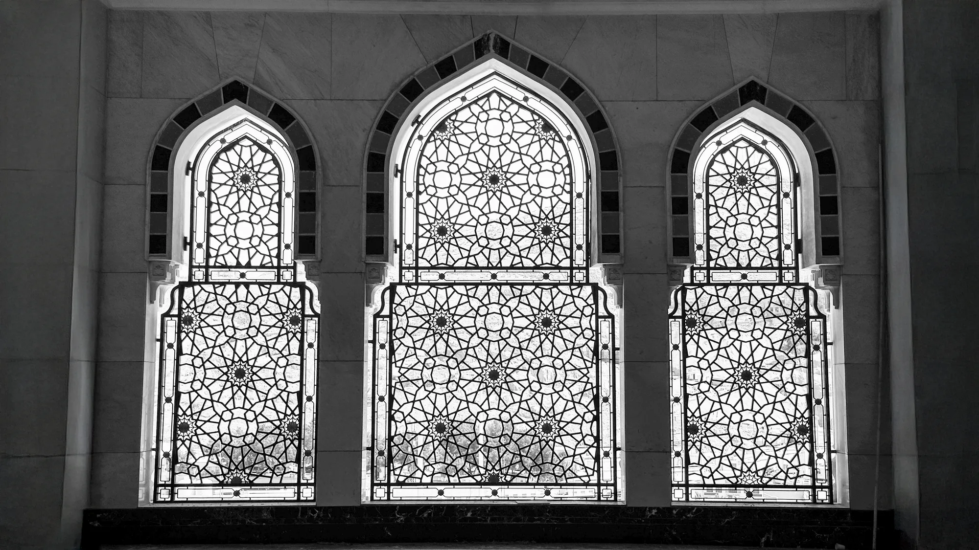 Black-and-white photograph of three geometric mashrabiyya windows in Al-Masjid Al-Haram, Mecca — intricate Islamic star-lattice pattern