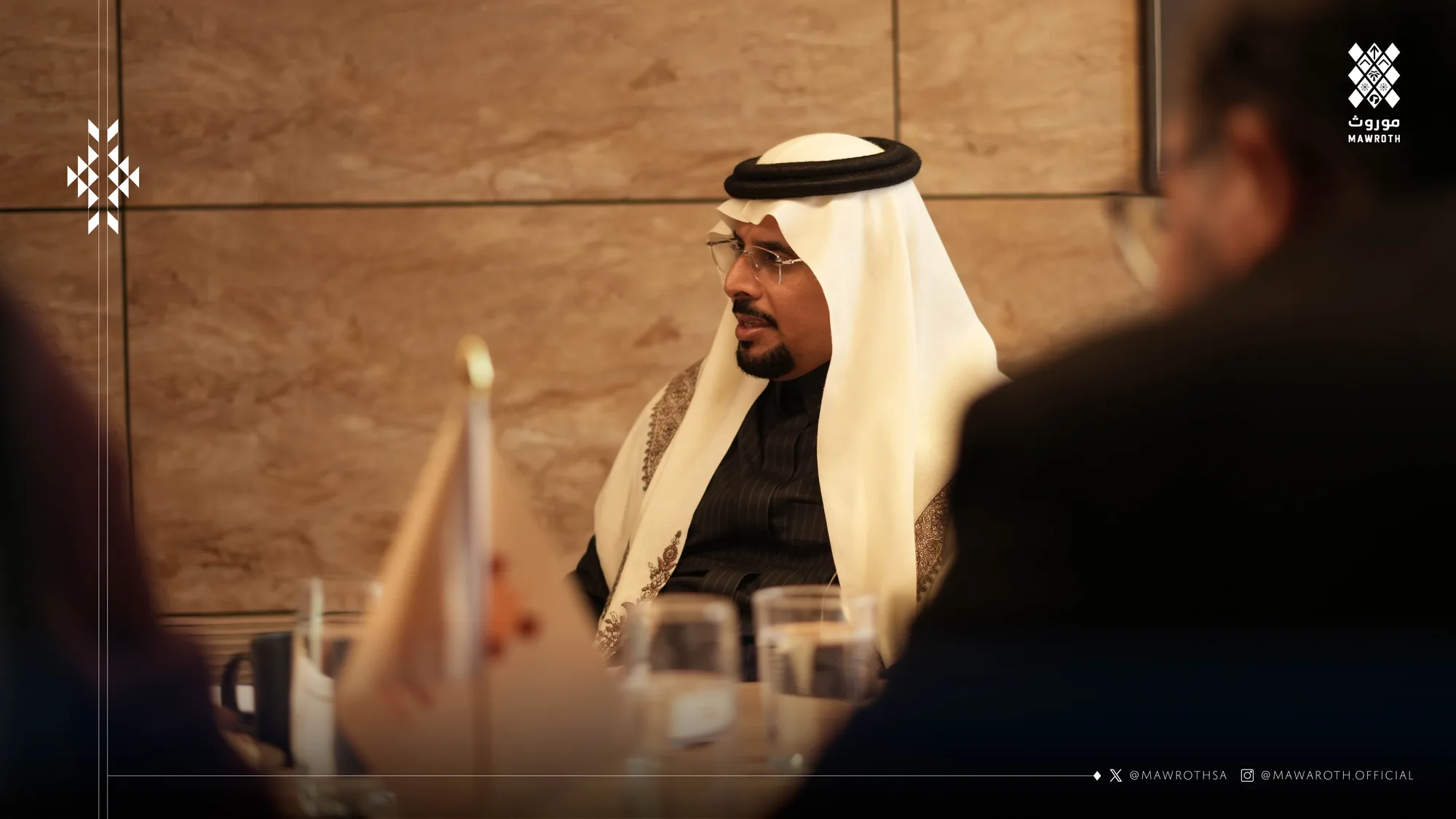 Senior Mawroth official addressing the meeting table, framed in close-up against the marble backdrop
