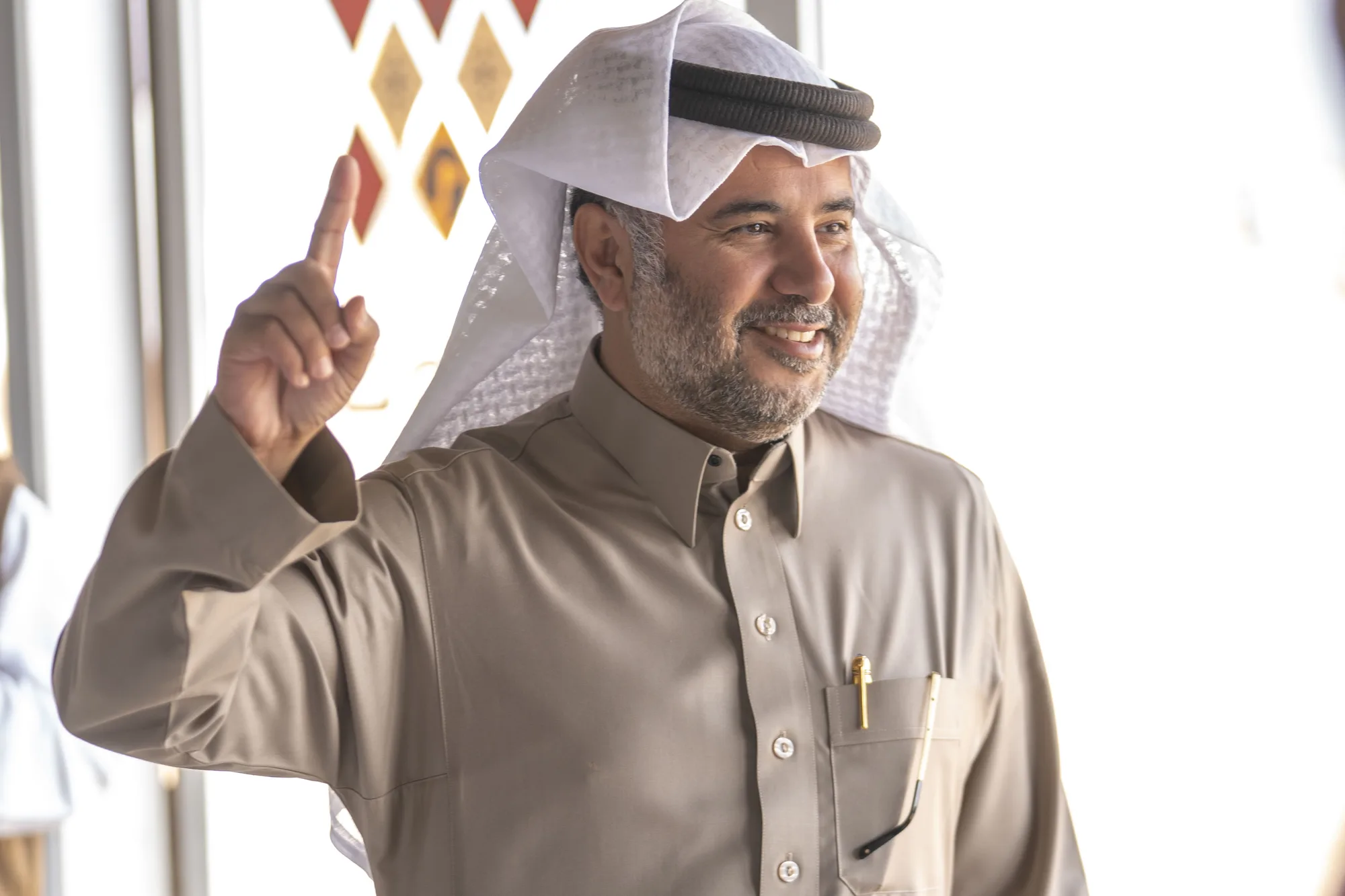 Senior official gesturing animatedly at the Taif Falcon Cup 2025 venue, diamond-motif heritage wall behind him