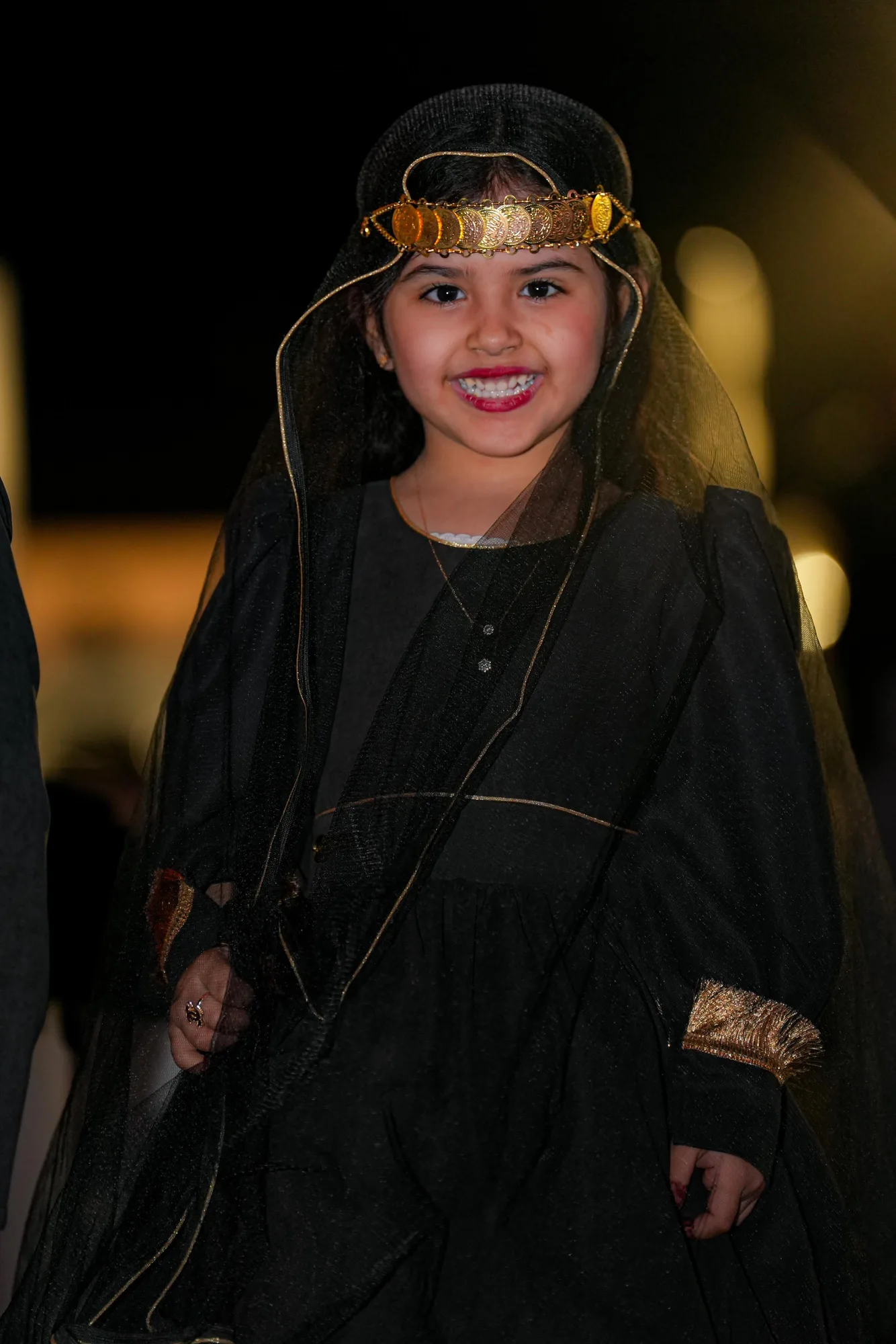 Young girl in traditional Saudi ceremonial dress with gold headdress smiling at the Taif Falcon Cup night gala