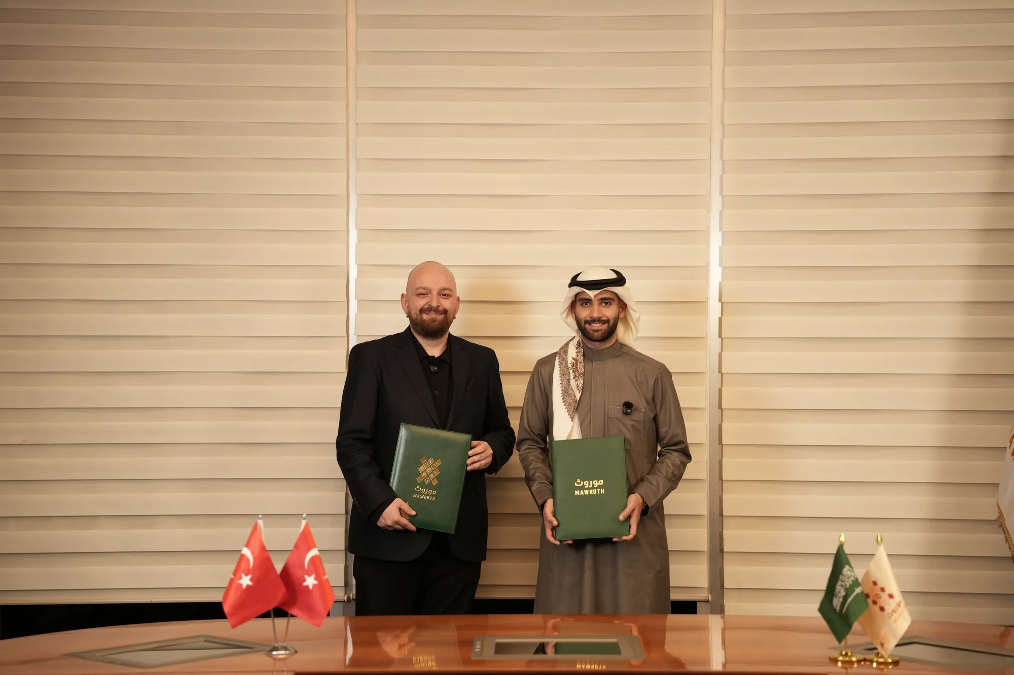 Mawroth and UNLOST representatives holding branded signing folders at the MOU ceremony, Turkish and Saudi flags on the desk